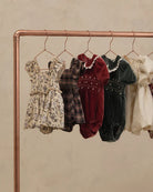 Five baby dresses and smock suits in various patterns and colors hanging on a copper-colored clothing rack against a textured beige wall.