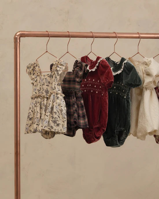 Five baby dresses and smock suits in various patterns and colors hanging on a copper-colored clothing rack against a textured beige wall.