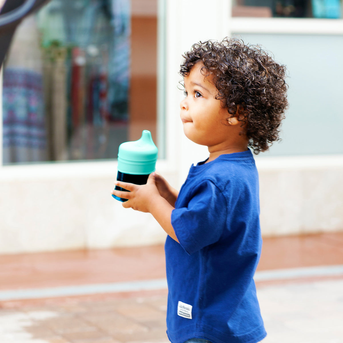 A young child with curly dark hair stands outdoors holding a teal and black sippy cup, wearing a blue short-sleeve shirt.