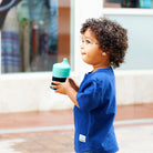 A young child with curly dark hair stands outdoors holding a teal and black sippy cup, wearing a blue short-sleeve shirt.