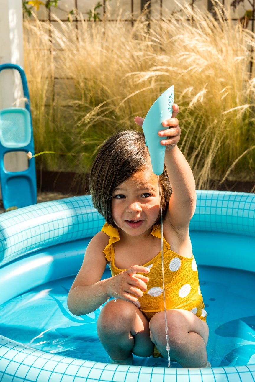 Child in a yellow swimsuit with white polka dots sitting in a blue inflatable pool.