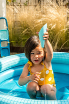 Child in a yellow swimsuit with white polka dots sitting in a blue inflatable pool.