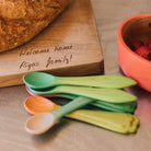 Colorful wooden spoons on a wooden surface with a bowl of strawberries and a cutting board with a message.