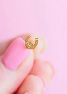 A close-up of a hand with pink nail polish holding a small gold horseshoe-shaped earring against a soft pink background.