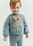 Child wearing a light blue sweatshirt with a teddy bear design on a plain background
