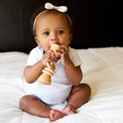 Baby sitting on a white blanket with a wooden toy, wearing a white outfit and headband.