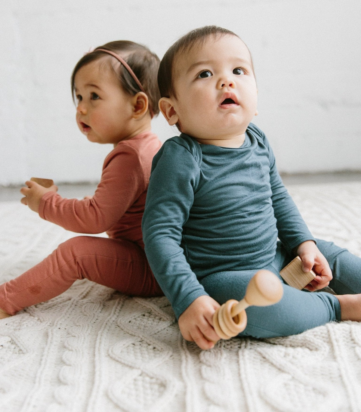 Two babies sitting on a textured surface, one in a pink outfit and the other in a blue outfit, playing with wooden toys.