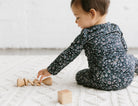 Baby in a floral onesie playing with wooden toys on a white blanket.