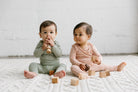 Two babies playing with wooden blocks on a white blanket.
