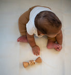 Baby in a brown outfit with a white bib playing with a wooden toy on a light surface.
