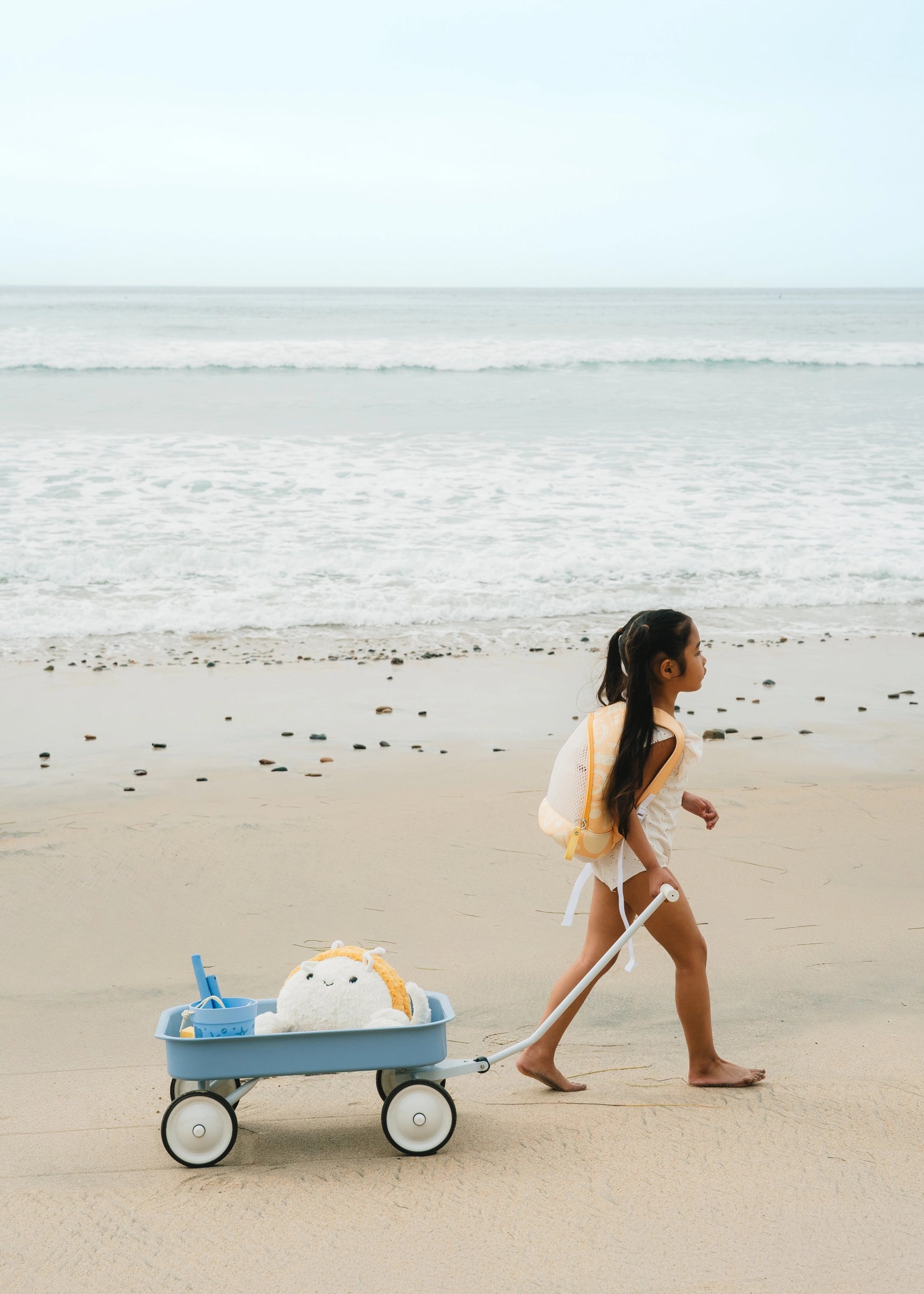 Woman walking on a beach with a blue cart pulling a plush toy