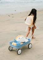 Child pushing a toy turtle in a blue wagon on a sandy beach.