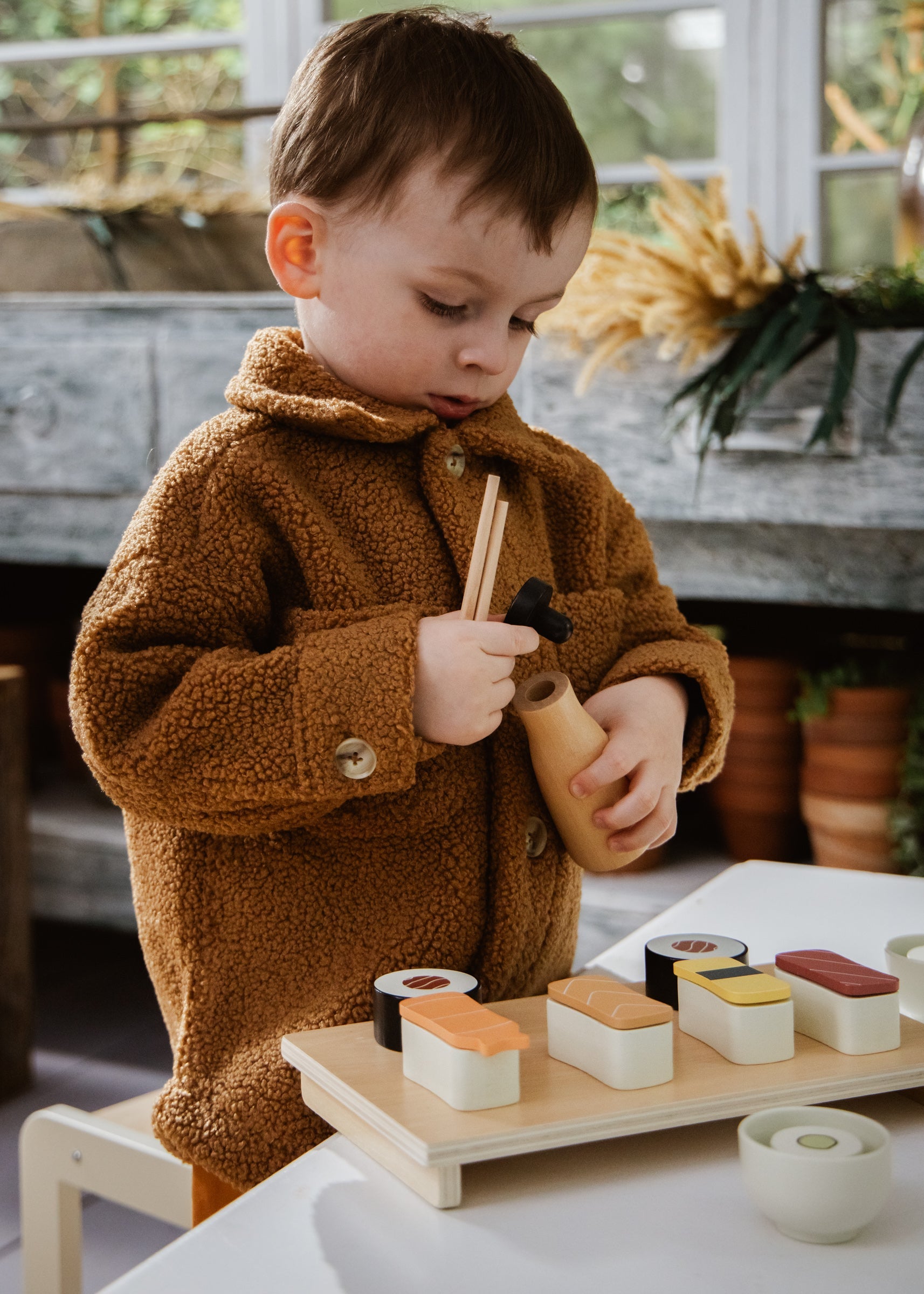 Child in a brown coat holding chopsticks over a tray of toy sushi.