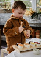 Child in a brown coat holding chopsticks over a tray of toy sushi.