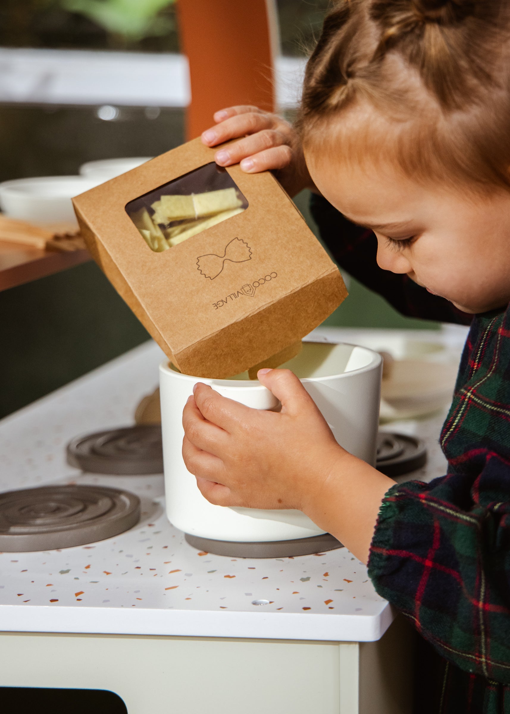 Child interacting with a small white appliance on a stove, with a cardboard box featuring an illustration of a bow tie.