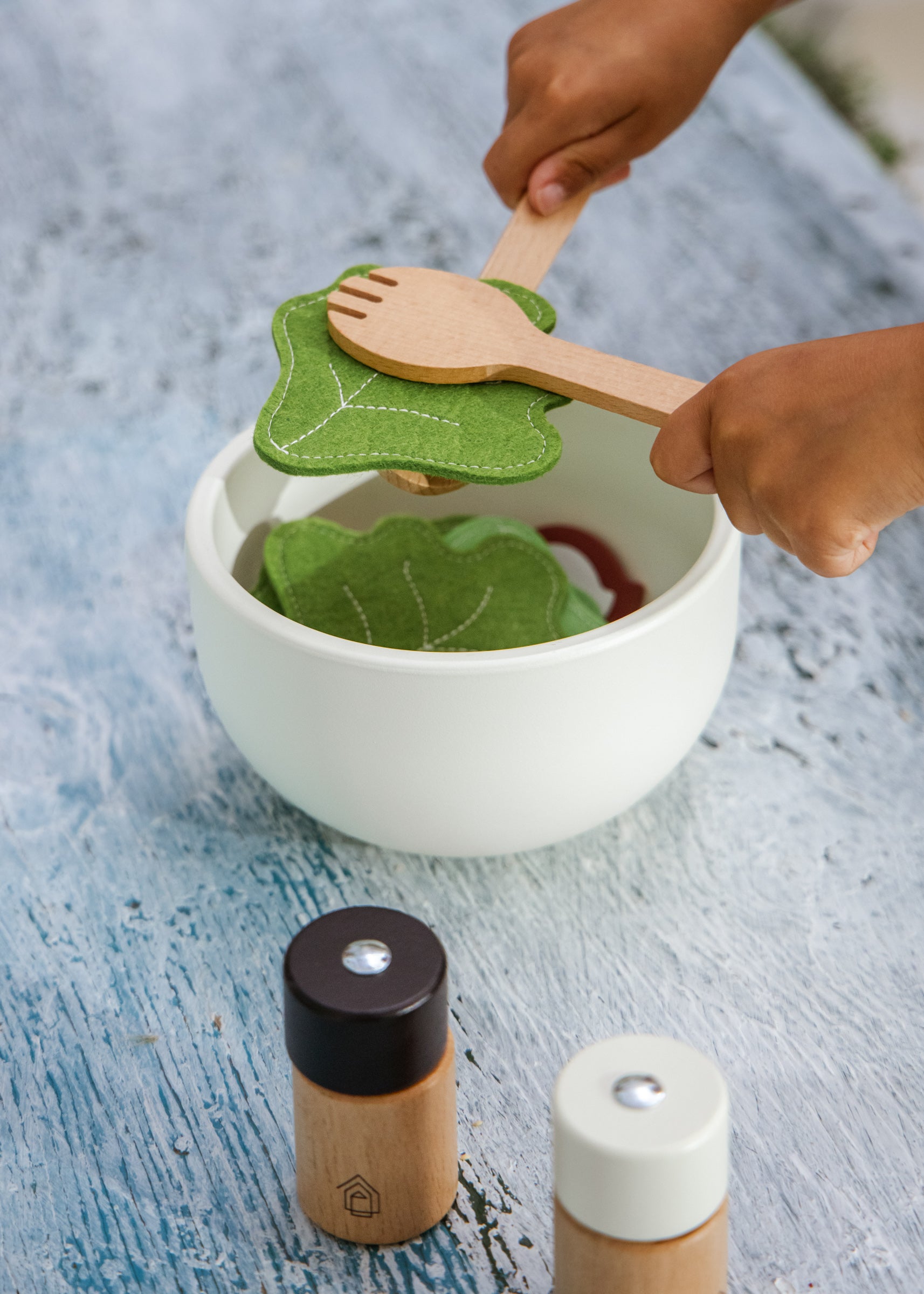 Children's play kitchen set with a white bowl, green leaves, and wooden utensils on a textured surface.