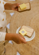 Hands holding a wooden tray with a slice of bread and a small round plate with a similar setup on a blurred background.
