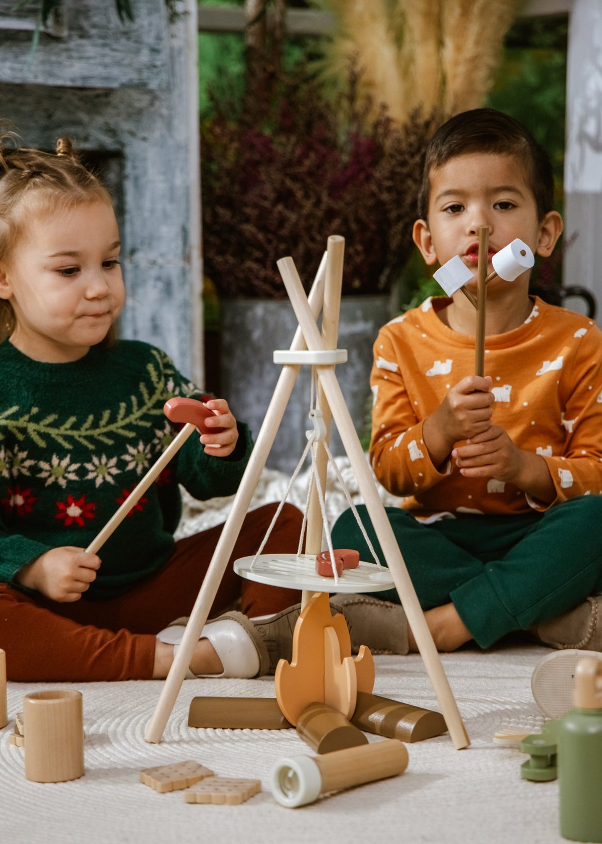 Two children playing with wooden toys including a teepee and campfire.