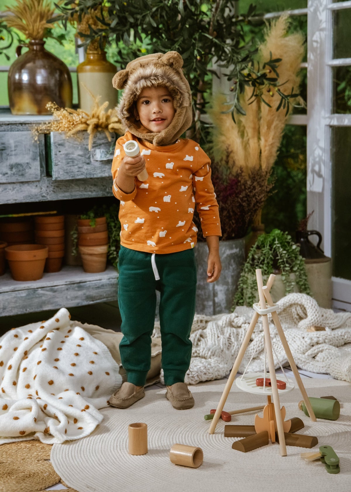 Child in orange hoodie and green pants standing in a room with plants and wooden toys.