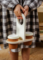 Person holding a wooden cup holder with three brown cups, wearing a checkered robe.