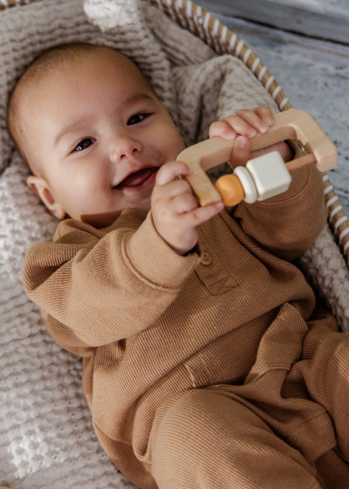 Baby in a brown outfit holding a wooden toy, sitting in a basket with a textured blanket.