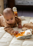 Baby in a brown outfit playing with a book and toys on a white blanket.