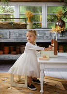 Young girl in a white dress standing next to a table with a wooden box in a rustic kitchen.