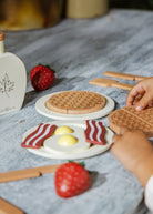 Wooden toy waffle with a child's hand reaching for it on a table with strawberries and a plate.