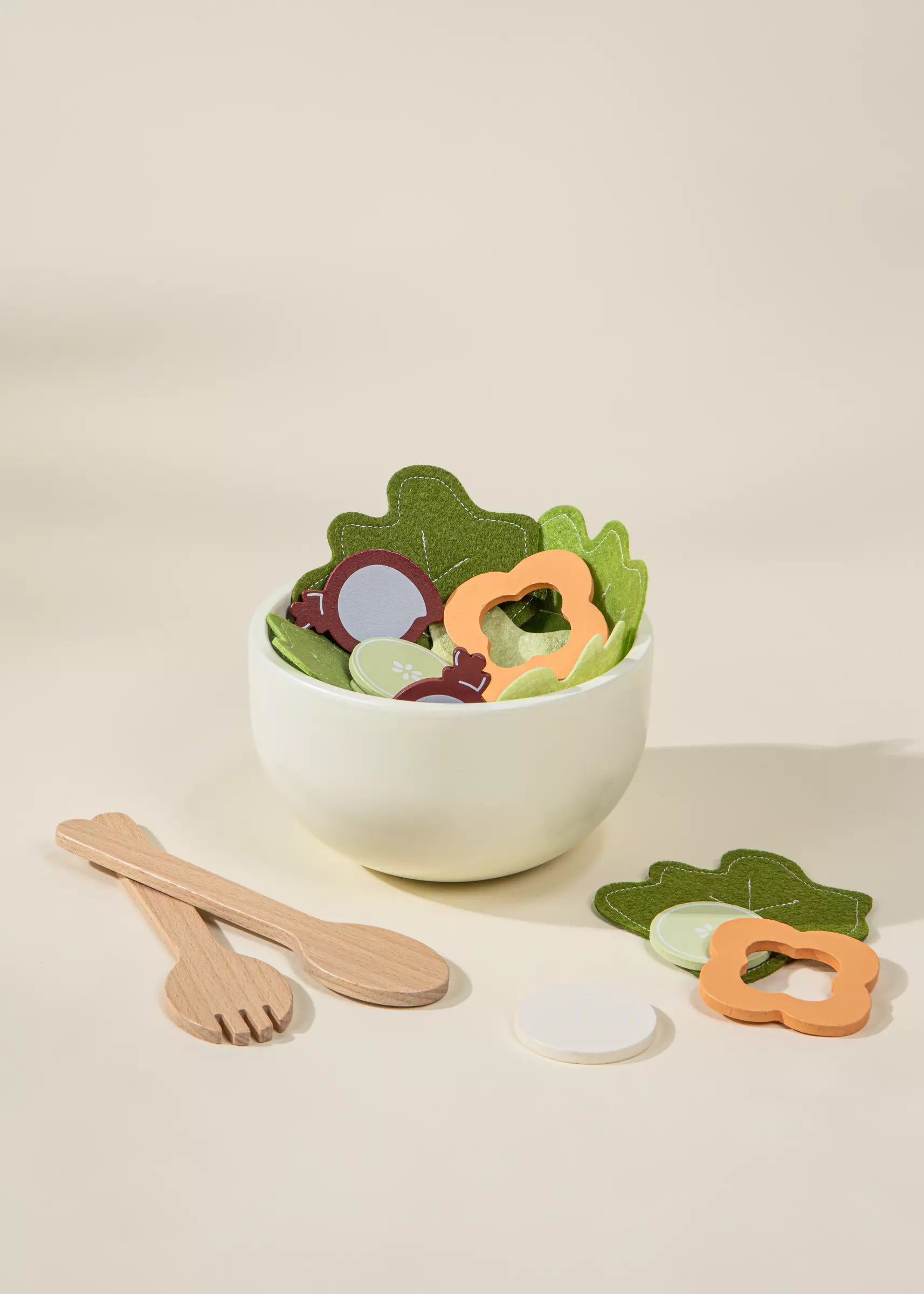 Children's toy salad set with a bowl, utensils, and colorful leaf-shaped pieces on a beige background.
