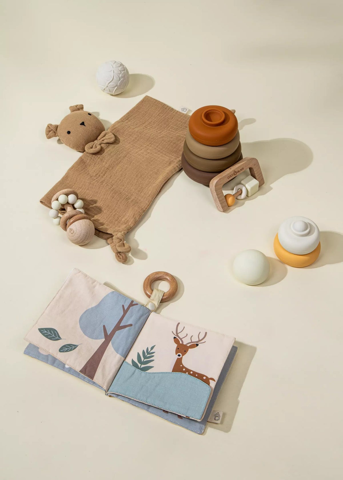 Collection of baby toys including a soft toy, wooden rings, and a fabric book with animal illustrations on a neutral background.