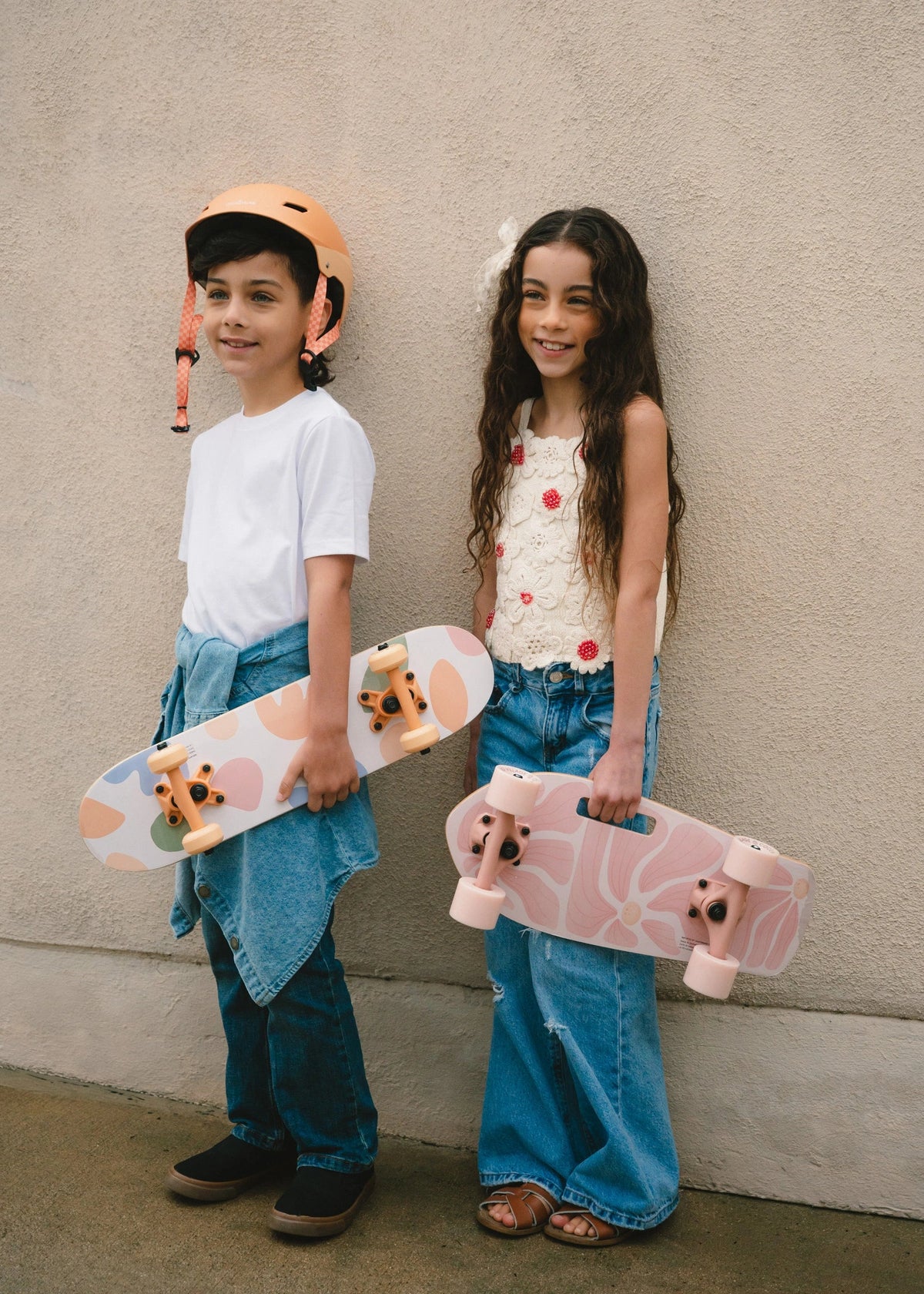 Two children holding skateboards against a plain wall.