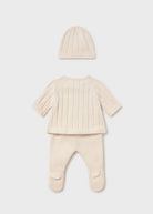 A three-piece baby knit outfit set in cream, featuring a short-sleeve footed sleeper, a matching beanie, and a ribbed long-sleeve top, laid out on a light gray background.