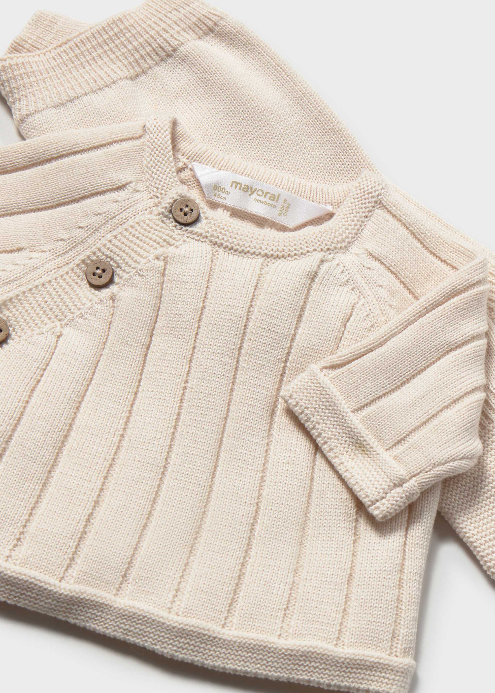 A close-up of a cream-colored ribbed knit baby cardigan with three wooden buttons, featuring a visible 'mayoral' brand label on the inside neck.