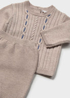 A close-up of a beige ribbed knit sweater with decorative blue embroidered details, shown alongside matching beige knit pants on a light gray background.