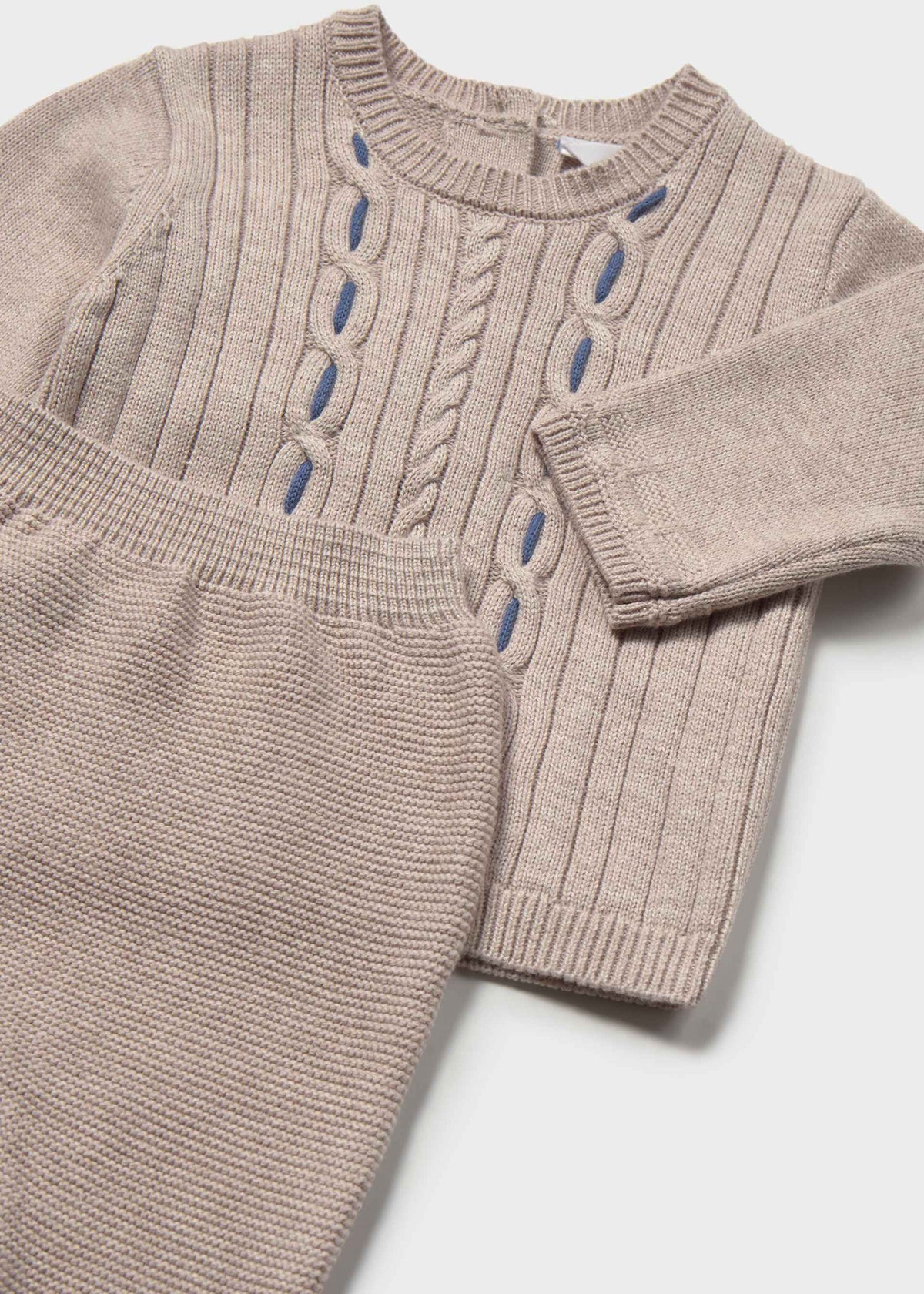 A close-up of a beige ribbed knit sweater with decorative blue embroidered details, shown alongside matching beige knit pants on a light gray background.