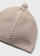 A close-up of a light taupe knit beanie hat featuring a vertical cable-knit pattern and a matching pom-pom on top, set against a clean white background.