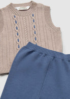 Knitted sleeveless top with cable pattern and blue pants on a light gray background