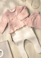 A baby clothing set laid flat on a light wood surface, including a pale pink knitted cardigan with pom-pom texture, cream ribbed footed pants, and a matching pink knit hat with a pom-pom. A small box is partially visible underneath.