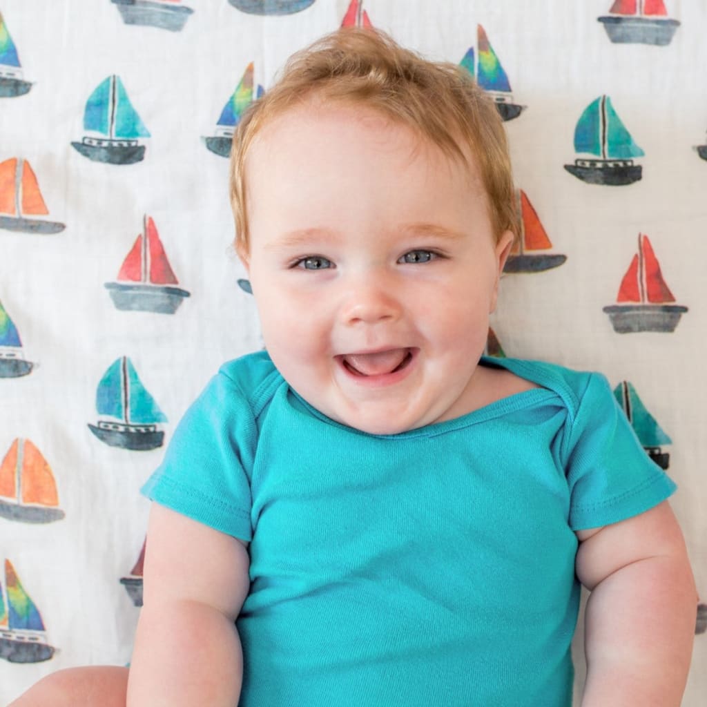 Baby in a teal onesie sitting against a background with sailboat patterns