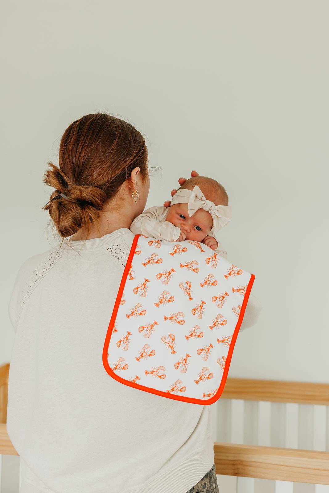 Woman holding a baby wrapped in a white blanket with orange patterns, standing against a light-colored wall.