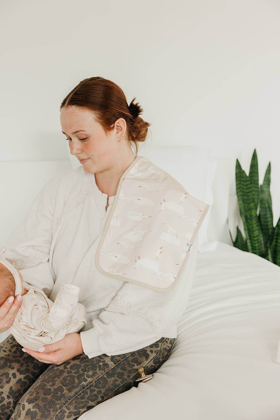 Woman sitting on a bed holding a baby wrapped in a blanket, with a plant in the background.