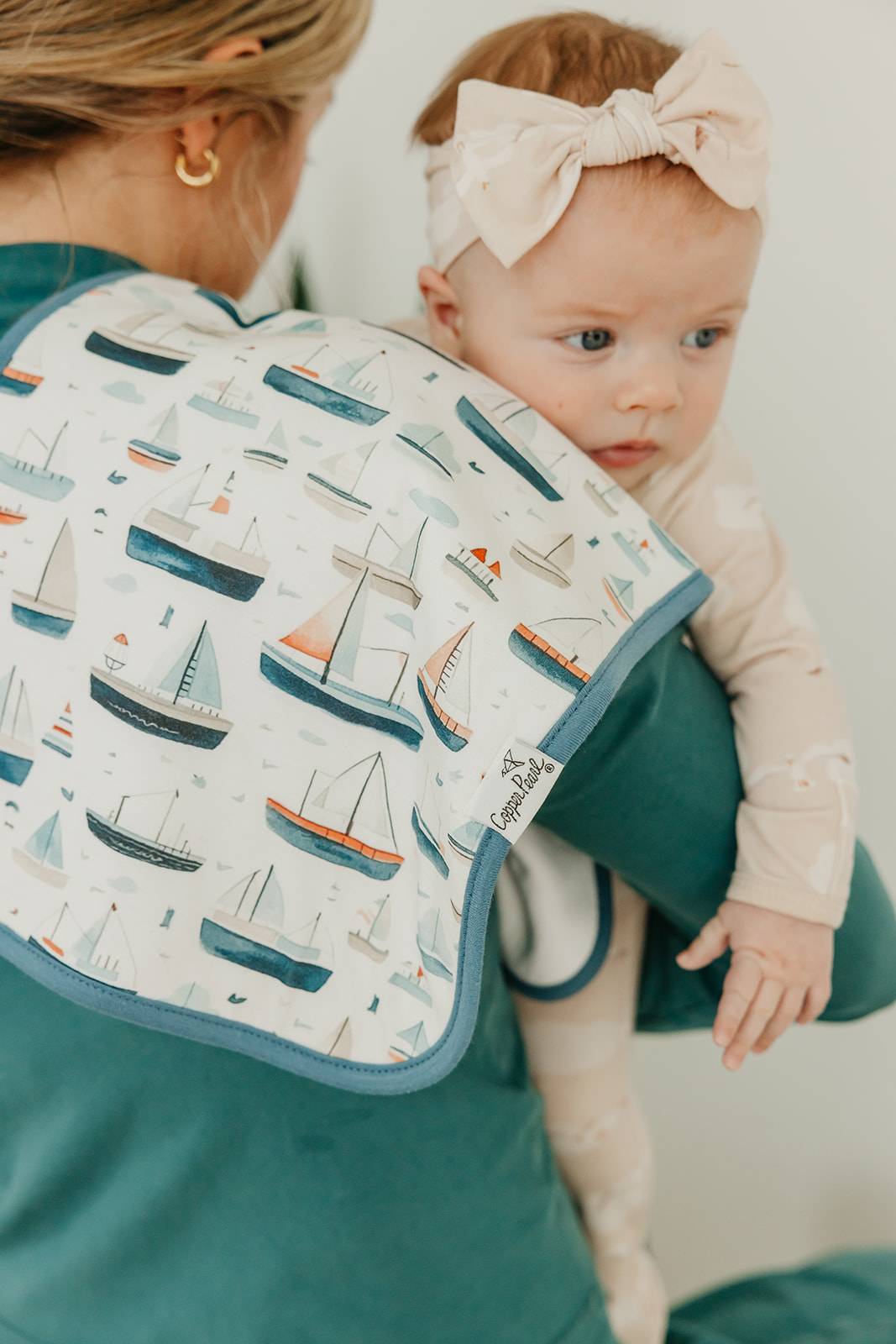 Baby wearing a bib with boat pattern and a headband, sitting next to an adult.