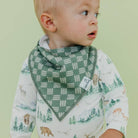 A baby wearing a green and white patterned bandana bib over a white long-sleeve shirt with a forest and animal print, set against a solid pale green background.