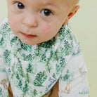 A close-up of a baby wearing a forest-print bib and matching top. The white fabric features green pine trees, brown foxes, and deer, with a visible 'Cappella' brand label on the bib.