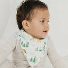 A baby wearing a white cotton bib with a woodland print of deer, trees, and birds, paired with a light heather gray long-sleeve shirt against a bright white background.