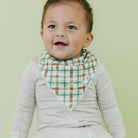 A smiling baby wearing a light gray long-sleeve romper and a green, brown, and white plaid bib, set against a solid pale green background.