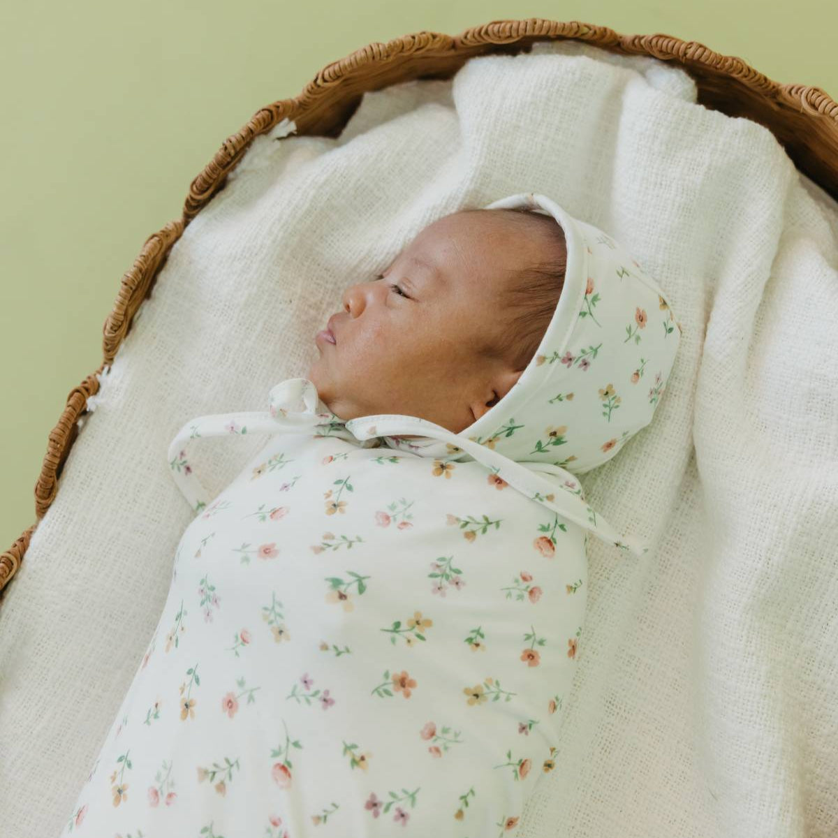 Newborn baby swaddled in a floral blanket with a green headband, lying in a woven basket against a light green background.