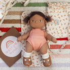 Toy doll in a pink outfit lying on a striped blanket with a rainbow card.