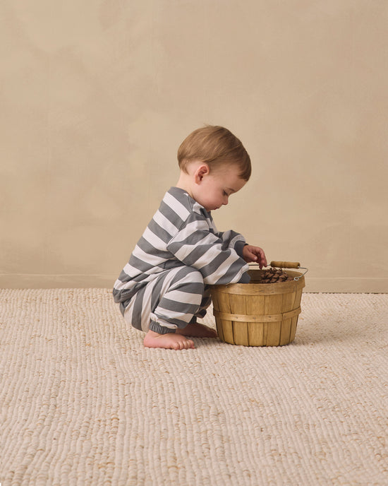 A baby wearing a gray and white striped long-sleeve romper crouches on a textured cream rug next to a small wooden bucket.