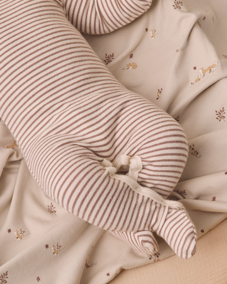 A close-up of a long, cylindrical nursing pillow with thin brown and beige stripes, nestled in a beige patterned blanket with small embroidered deer.